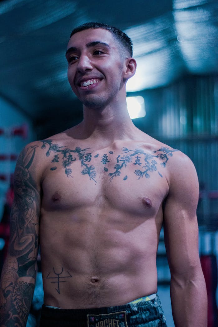 Portrait of a smiling muscled man with tattoos standing topless in a gym.
